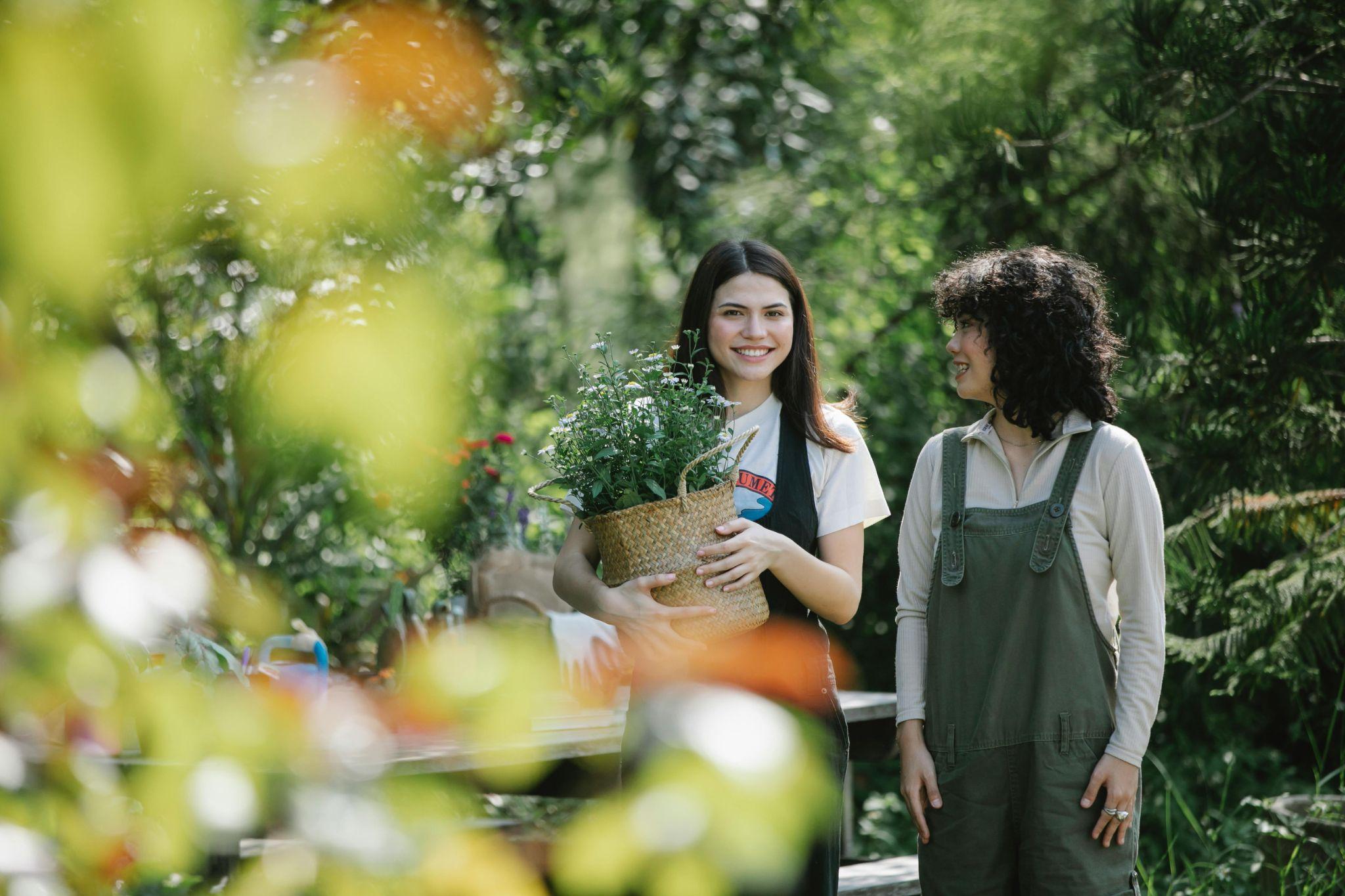 two women walking in a green garden carrying plants