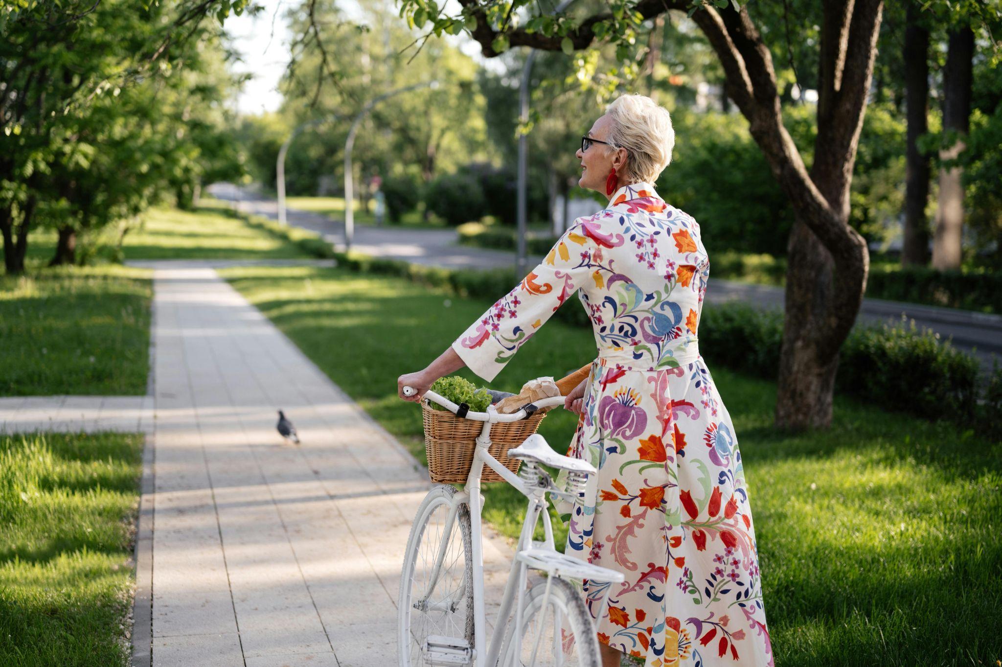 woman in a floral dress walking a bicycle on a tree-lined path