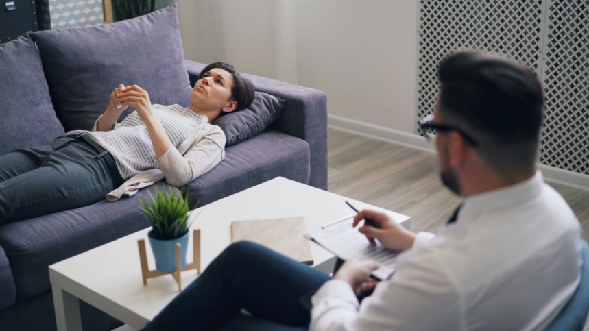 Person lying on a sofa during a mental health consultation while a clinician takes notes in a quiet, professional setting