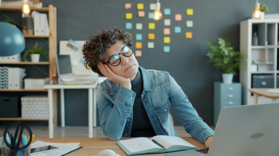 Person sitting at a desk, looking tired and unfocused while working on a laptop with notes and books nearby