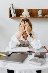 Woman sitting at a table with papers and laptop, covering her face in frustration