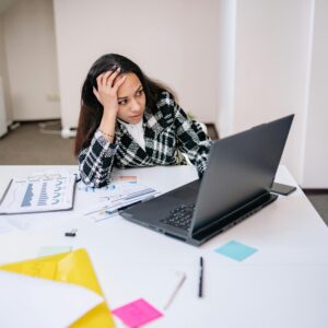 Woman sitting at a desk with a laptop, holding her head while reviewing papers and charts